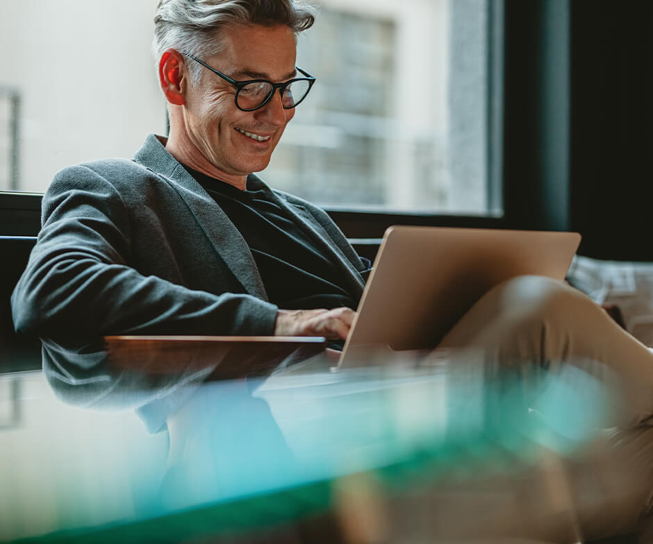 Auf einer Couch sitzende Person mit Brille und Business-Anzug, die auf einen Laptop blickt und lächelt