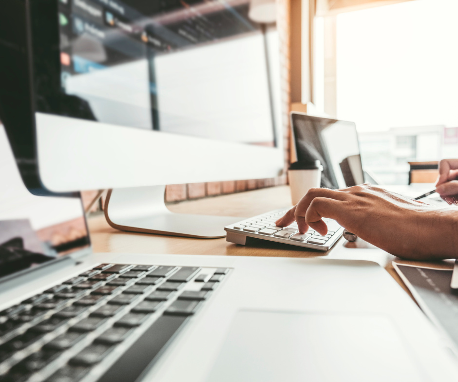 una persona escribiendo en el teclado delante de un monitor externo y una taza de café en la mesa