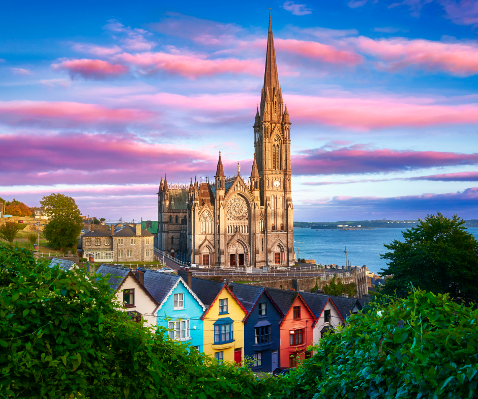 Un cielo colorido y salpicado de nubes hace de fondo de un barrio de Cork con casas coloridas y el campanario de una iglesia.